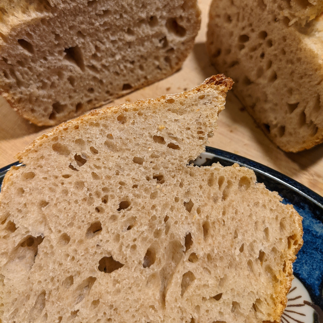 A slice of sourdough bread on a plate with the loaf in the background, focus is on the crumb of the bread