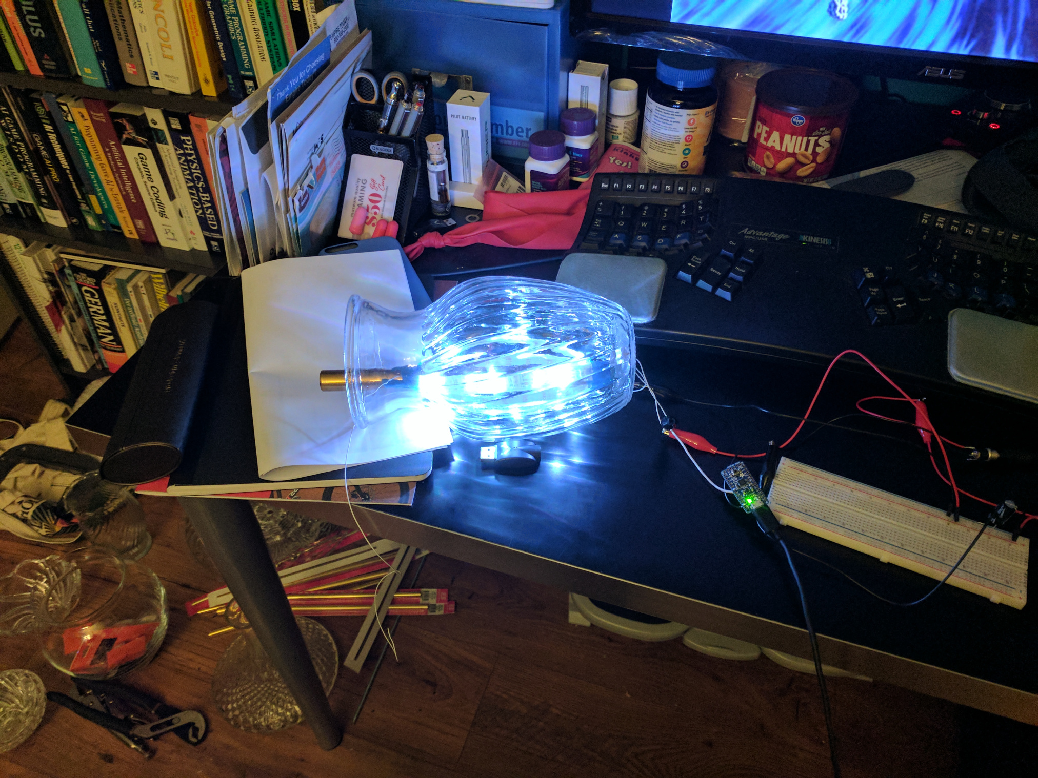 A clear, textured vase with LEDs shining inside, laying on its side on a messy desk with some electronics.