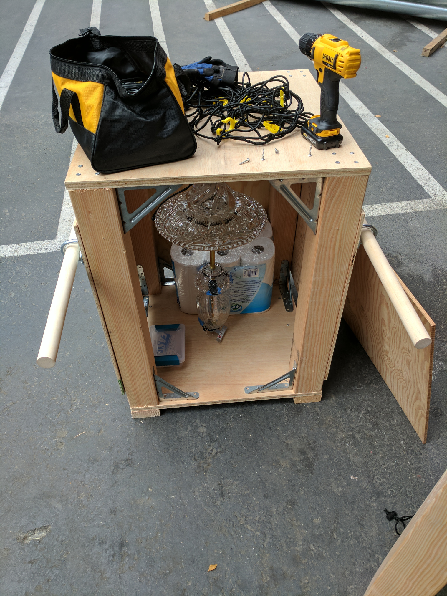 The crate, with 3 sides closed, sitting in a parking lot.  The lantern hangs inside, braced by a 12 pack of toilet paper. Tools sit on top of the crate.