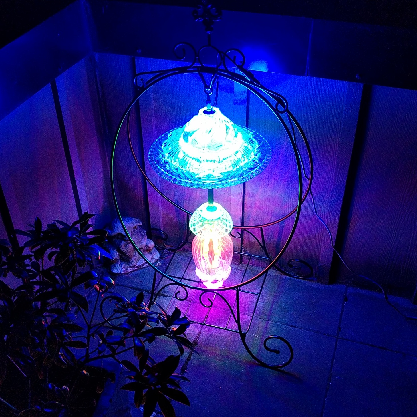 The lantern, brightly glowing blue and pink, hanging in a fancy, circular, wrought iron plant stand on a condo balcony, near a plant, at night.