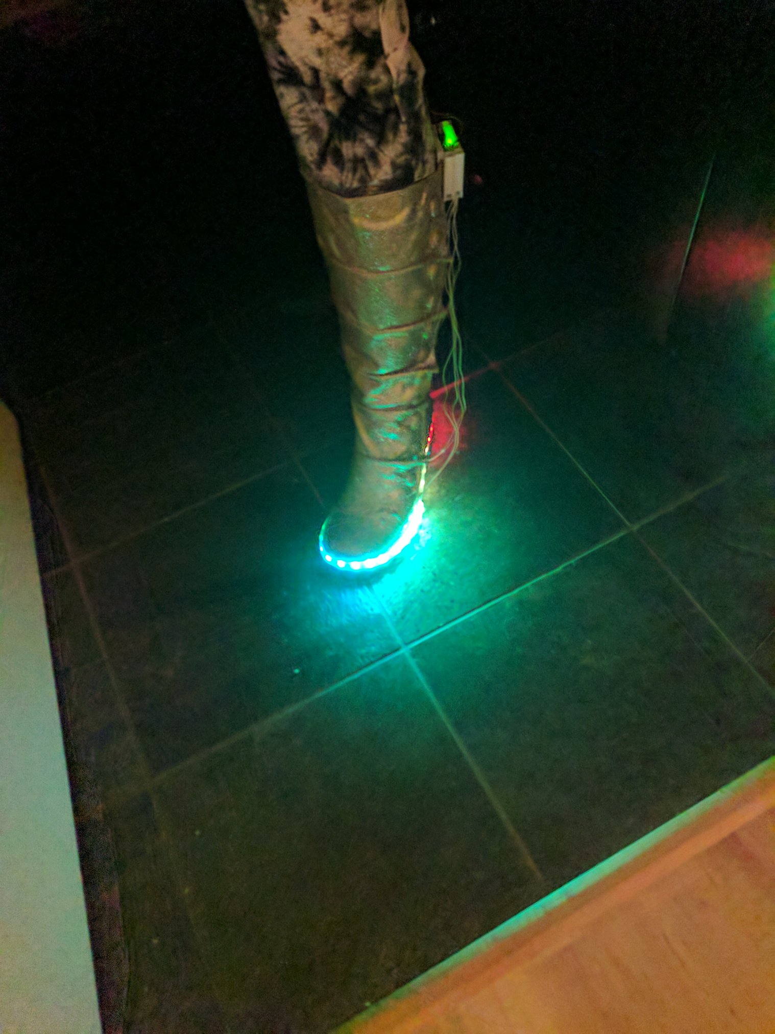 A leg, wearing a gold glitter covered calf high boot with LEDs along the base and a home made electronics enclosure on the back.  Wires hang loose. The LEDs glow blue.