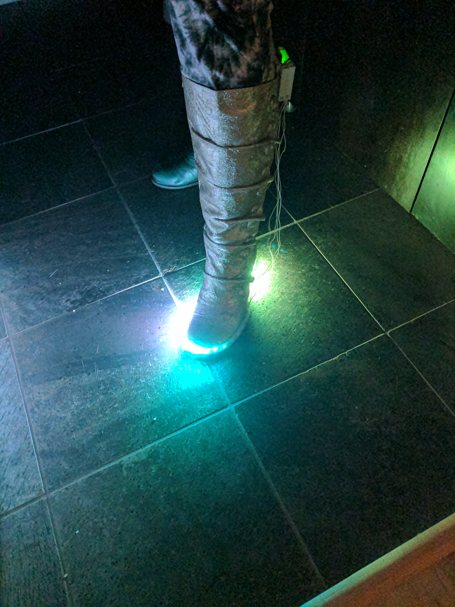 A leg, wearing a gold glitter covered calf high boot with LEDs along the base and a home made electronics enclosure on the back.  Wires hang loose. The LEDs glow on one side, part way through an animation.