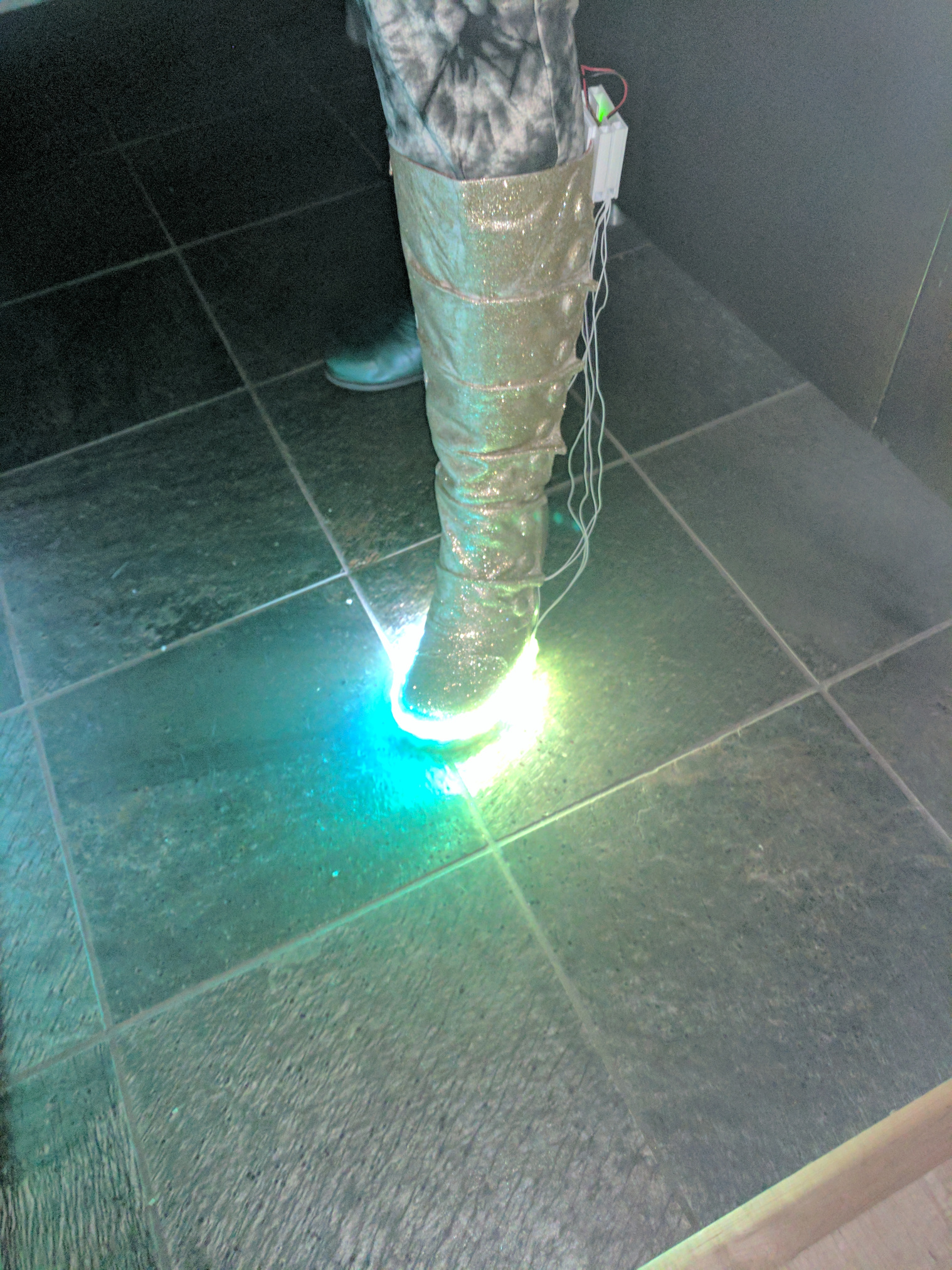 A leg, wearing a gold glitter covered calf high boot with LEDs along the base and a home made electronics enclosure on the back.  Wires hang loose. The LEDs glow blue and green.
