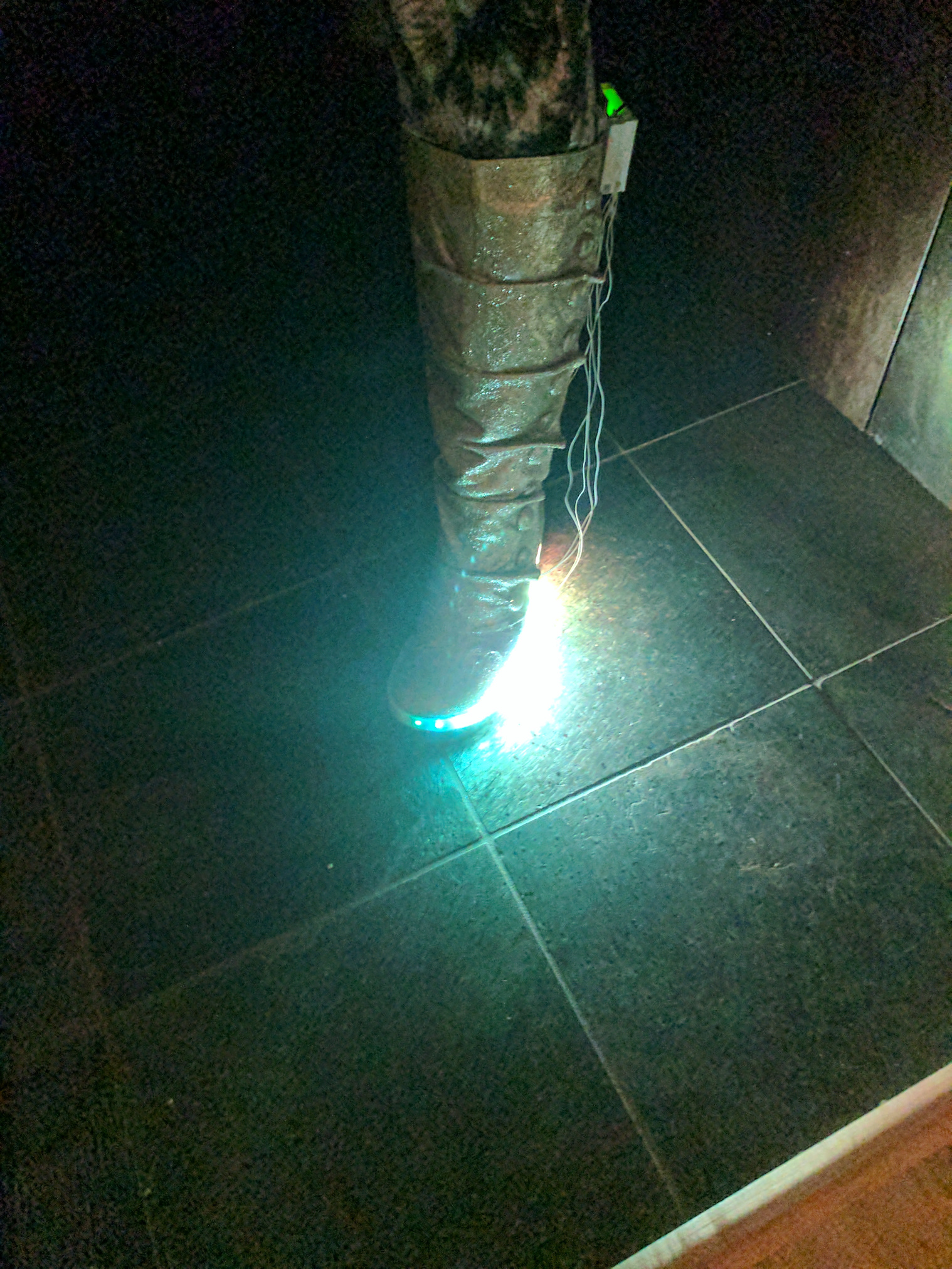 A leg, wearing a gold glitter covered calf high boot with LEDs along the base and a home made electronics enclosure on the back.  Wires hang loose. The LEDs glow white.