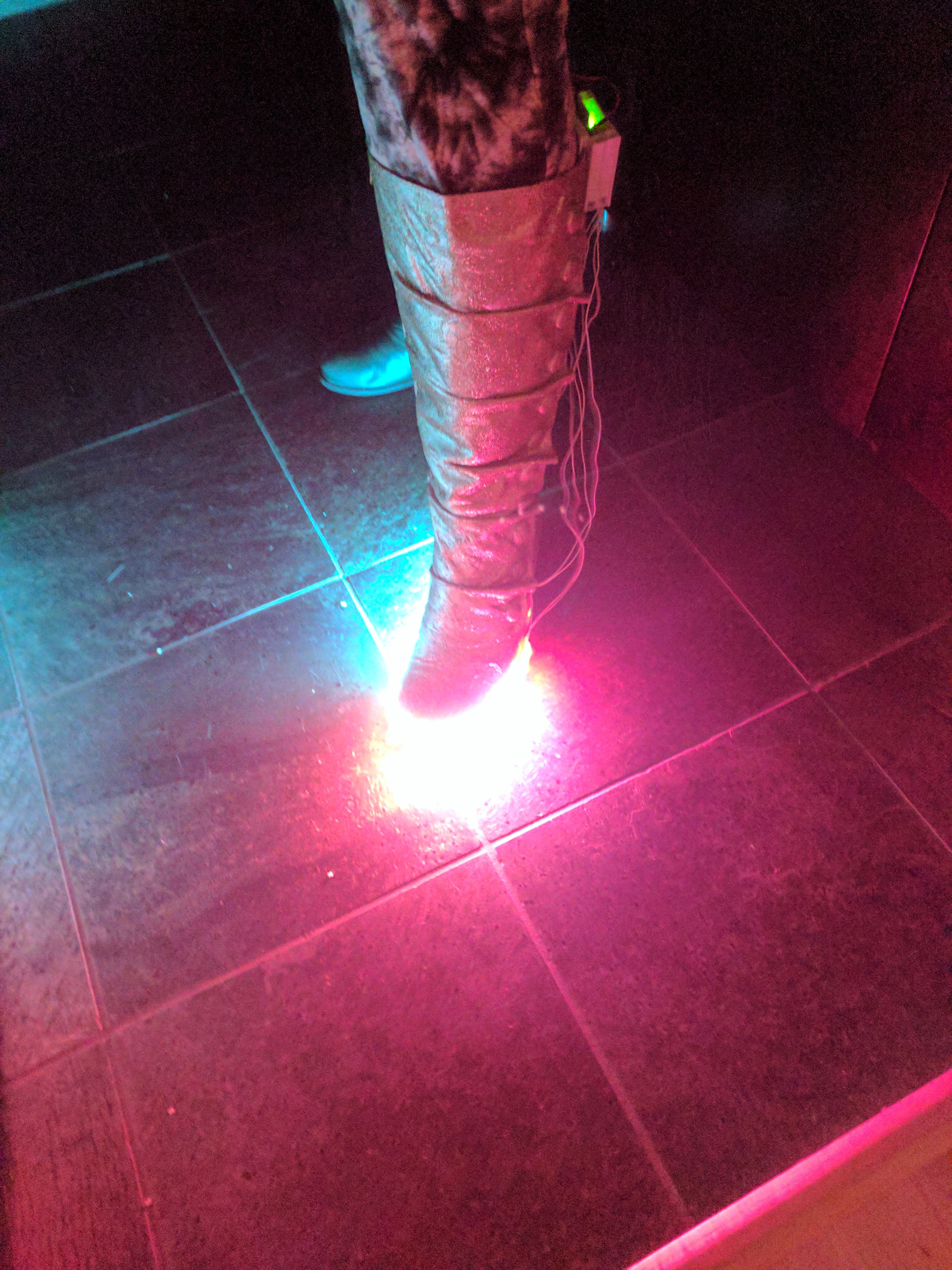 A leg, wearing a gold glitter covered calf high boot with LEDs along the base and a home made electronics enclosure on the back.  Wires hang loose.  The LEDs glow pink.