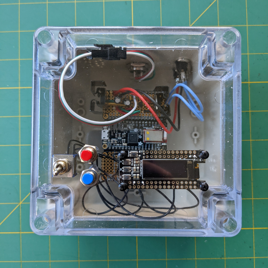 A plastic project box with a clear face. A switch, a red button, a blue button, and a screen are mounted to the front of the box