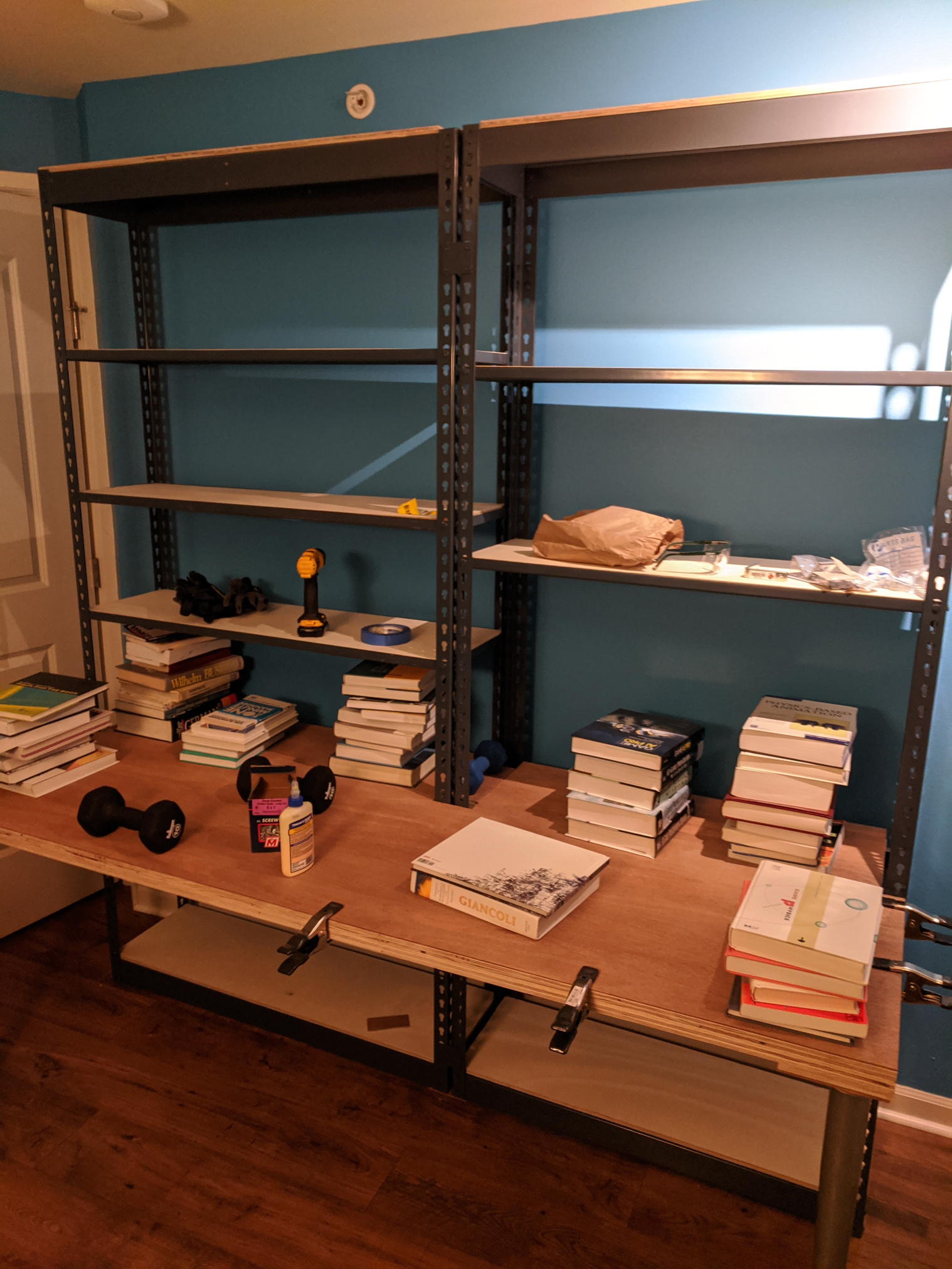 Two connected metal rivet shelving units with a desk top mounted to the front. Desk is clamped and weighted down with books.